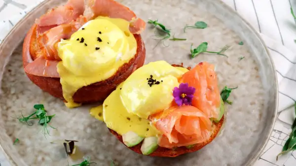 Dos deliciosos huevos benedictinos, uno con jamón y otro con salmón ahumado y aguacate, servidos sobre pan tostado con salsa holandesa.