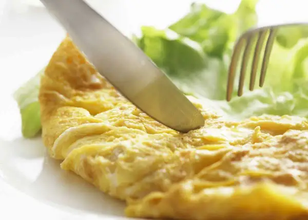 Un cuchillo corta una dorada tortilla francesa junto a hojas de lechuga fresca.
