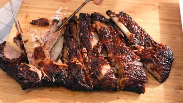 Costillas a la barbacoa jugosas y tiernas, cortadas en una tabla de madera. La carne se desprende del hueso, cubierta de salsa.