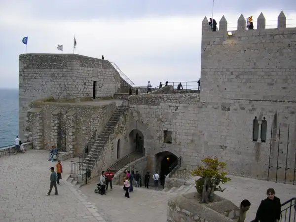 El castillo de Peñíscola se alza imponente sobre el mar. Turistas pasean por sus patios de piedra, admirando la arquitectura histórica.