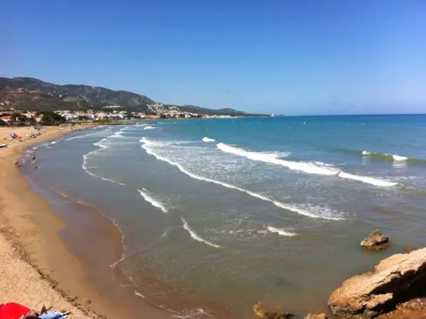 La **playa romana alcossebre** se extiende con arena dorada y aguas turquesas bajo un cielo azul. Olas suaves rompen en la orilla, mientras la costa se dibuja con casas y montañas al fondo.