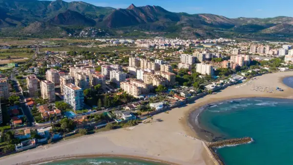 Vista aérea del casco antiguo de Benicàssim pueblo, con edificios residenciales, playas de arena y el mar Mediterráneo. Montañas verdes al fondo.