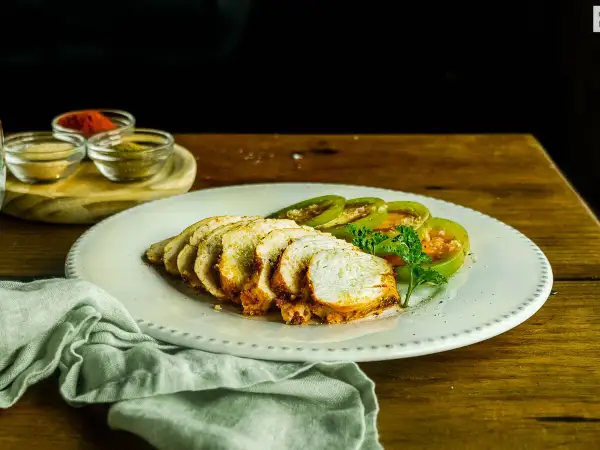 Pechugas de pollo al horno, jugosas y doradas, servidas con rodajas de tomate verde y perejil fresco. Especias en pequeños cuencos al fondo.
