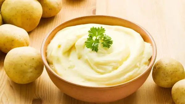 Un cremoso pure de patatas, decorado con perejil, junto a patatas frescas sobre una mesa de madera.