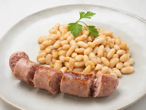 Un plato de alubias blancas tiernas y jugosas salchichas, decorado con una ramita de perejil fresco.